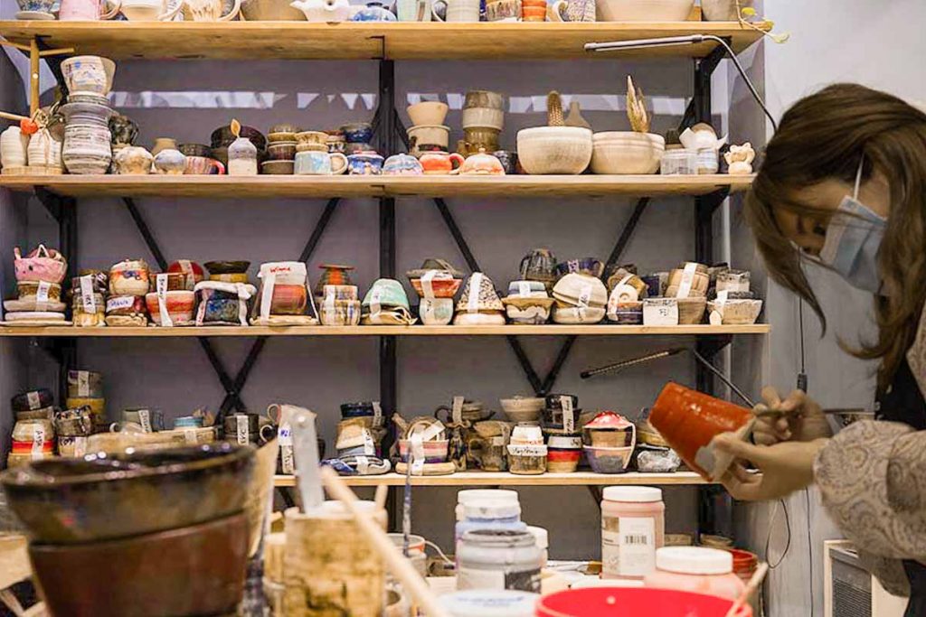 Woman paints ceramic with shelf of pottery behind her at Mudheytong Gallery - Things To Do In Hong Kong, Sham Shui Po