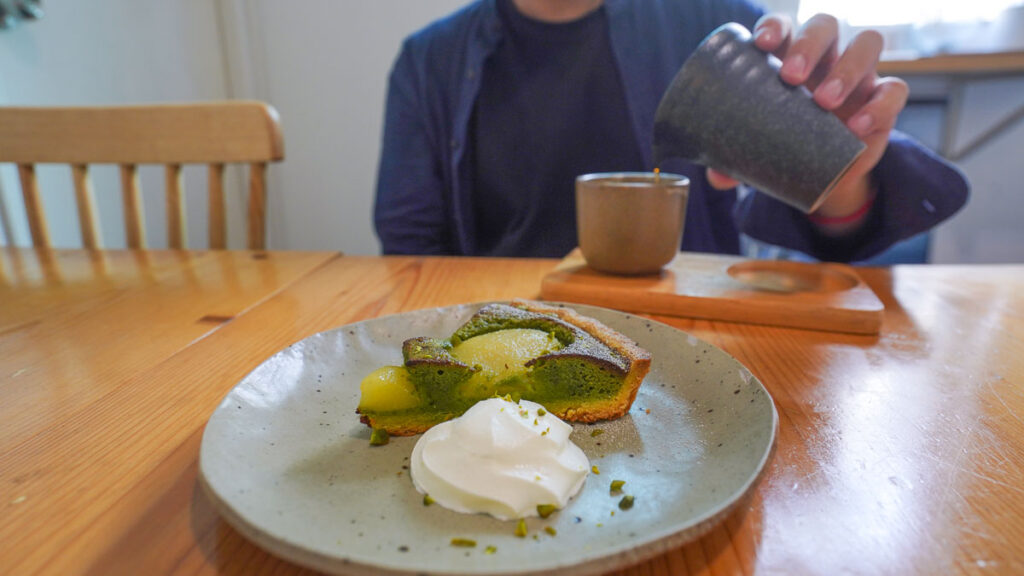 Poached Pear Pistachios Tart in foreground, person pouring out filter coffee in the background
