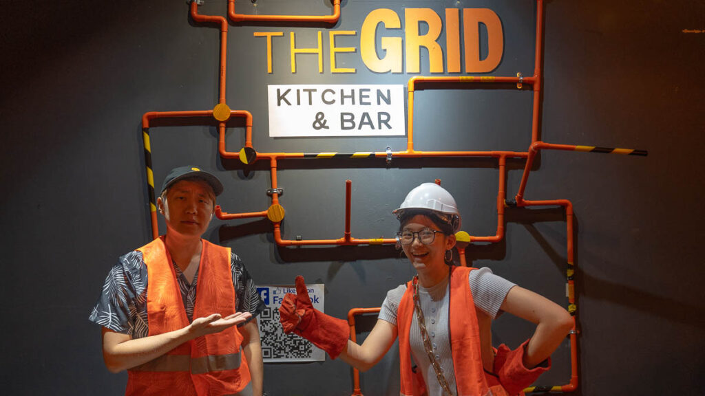 Two people posing in front of the Grid deco, wearing orange construction jackets - Skudai Food Guide