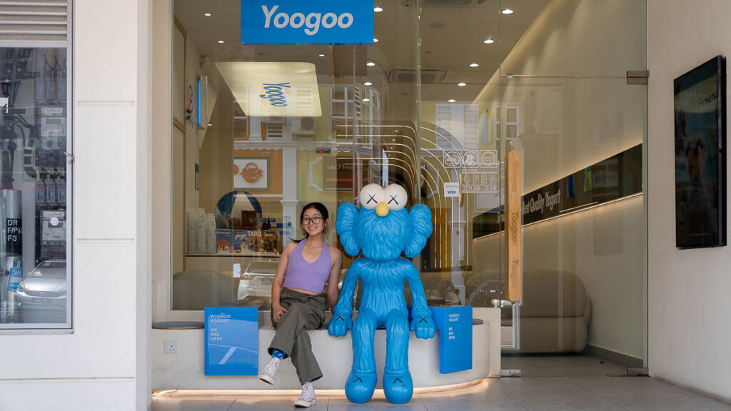 Person sitting beside a KAWS mascot at the storefront of Yogurt Yoogoo Mount Austin